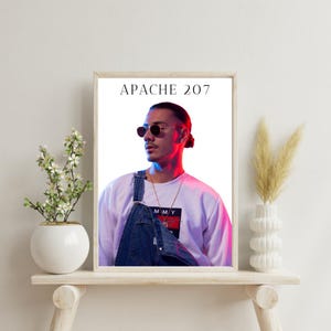Könnte beinhalten: Ein weißer gerahmter Poster mit dem Text "APACHE 207" in Schwarz. Der Poster zeigt ein Foto eines Mannes mit Sonnenbrille, einem weißen Sweatshirt und einer blauen Latzhose. Der Mann schaut nach links und hat ein rotes und blaues Licht, das auf ihn scheint.