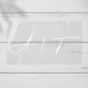 May include: A clear acrylic sign with the white script letters "A + T" on it. The sign is set against a white wooden background with horizontal planks. The image has a soft, natural lighting.