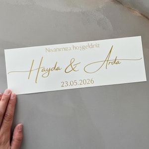 Puede incluir: Una tarjeta rectangular blanca con texto dorado. El texto dice "Nisanımıza hoşgeldiniz" encima de "Hayda & Arda" y la fecha "23.05.2026". La tarjeta está sobre una superficie gris.