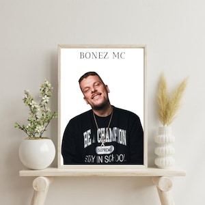 Könnte beinhalten: Ein weiß gerahmtes Poster mit dem Text "BONEZ MC" oben. Ein Mann mit kurzen braunen Haaren trägt einen schwarzen Pullover mit dem Text "BE A CHAMPION" und "STAY IN SCHOOL" in Weiß. Der Pullover hat auch ein kleines weißes Logo mit der Aufschrift "SUPREME".