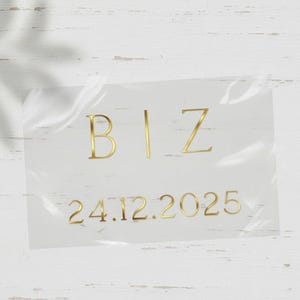 Könnte beinhalten: Ein transparenter Aufkleber mit goldfarbenen Buchstaben. Das Wort "BIZ" steht oben, darunter das Datum "24.12.2025". Der Aufkleber ist leicht zerknittert und liegt auf einer weißen, strukturierten Oberfläche.