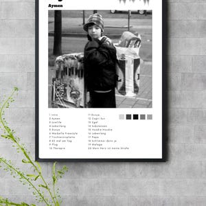 Könnte beinhalten: Ein gerahmtes Schwarz-Weiß-Poster mit dem Titel "Aymen" und einem Foto einer Person mit Hut. Unter dem Bild befindet sich eine Liste von Songtiteln. Das Poster hängt an einer Wand.