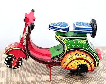 May include: A hand-painted wooden scooter in vibrant colors. The scooter features a red body with intricate gold and white detailing, a green seat, and yellow and orange accents. The wheels are black with white polka dots.