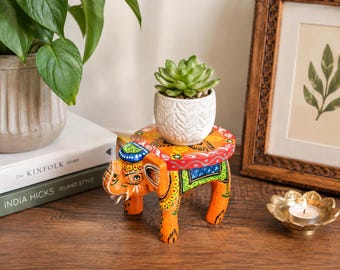 May include: A hand-painted wooden elephant stool featuring vibrant orange, blue, green, and yellow floral patterns. The elephant has a flat, circular top surface that supports a small, white, textured ceramic pot containing a green succulent plant.