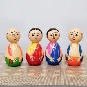 May include: Four wooden dolls, each with a different colored outfit. The dolls are painted with simple facial features and are wearing traditional Indian clothing. The dolls are all standing on a brown surface.