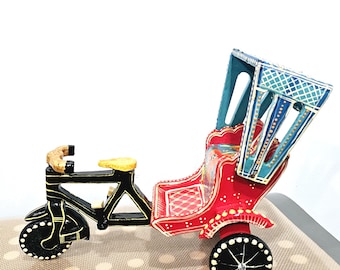 May include: A decorative, hand-painted rickshaw model. The miniature vehicle features a black frame, red passenger seat, and a blue canopy. The wheels are black with white accents, and the seat is yellow. The rickshaw is a charming piece of decor.