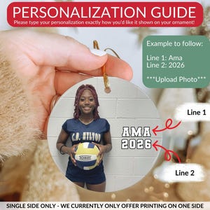 May include: A round white ornament featuring a printed photograph of a person in a navy blue athletic uniform holding a volleyball. The text AMA is printed on line one and 2026 is printed on line two. The ornament includes a gold hanging string.