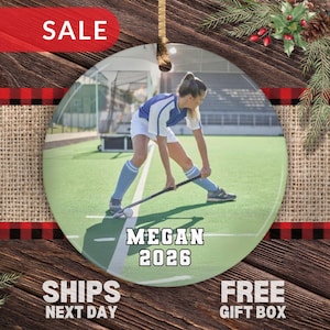 May include: A round ceramic ornament featuring a photograph of a field hockey player in a blue and white uniform on a green field. The text MEGAN and 2026 is printed in white with a black outline at the bottom of the circular ornament.