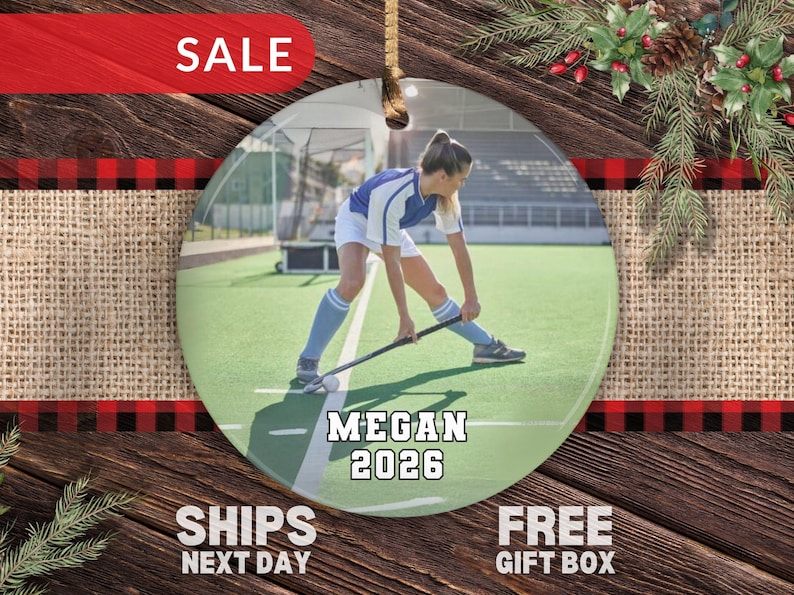 May include: A round ceramic ornament featuring a photograph of a field hockey player in a blue and white uniform on a green field. The text MEGAN and 2026 is printed in white with a black outline at the bottom of the circular ornament.