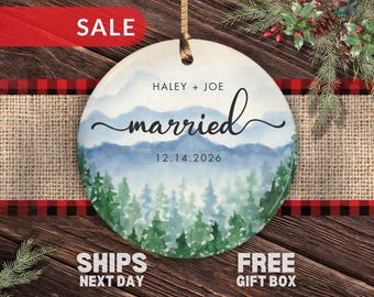 May include: A round ceramic ornament featuring a watercolor mountain and forest landscape. The text reads HALEY + JOE, married, and 12.14.2026. The ornament is suspended by a gold ribbon and displays a serene blue and green nature scene.