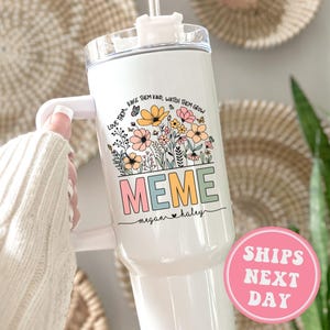 May include: White stainless steel tumbler with a floral design and the word "MEME" in colorful letters. The text "LOVE THEM, RAISE THEM KIND, WATCH THEM GROW" is written above the word "MEME". The names "megan & haley" are written below the word "MEME".
