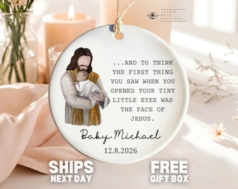 May include: A round white ceramic ornament featuring an illustration of Jesus holding a baby. The text reads: ...AND TO THINK THE FIRST THING YOU SAW WHEN YOU OPENED YOUR TINY LITTLE EYES WAS THE FACE OF JESUS. Baby Michael 12.8.2026. It has a twine hanger.