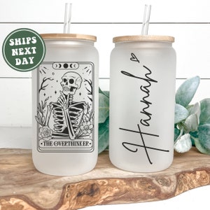 May include: Two frosted glass tumblers with bamboo lids and straws. One tumbler features a black and white illustration of a skeleton with the text "The Overthinker". The other tumbler has the name "Hannah" written in black cursive.