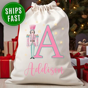 May include: Cream-colored drawstring gift bag with a pink letter "A" and the name "Addison" in pink script. A nutcracker soldier and gold stars decorate the bag. A green circle with the words "SHIPS FAST" is in the upper left corner.