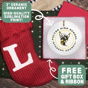 Op de afbeelding: Een rond wit keramieken ornament van 7,6 cm met een geel-zwarte horzelafbeelding en de tekst Fulton High School Student Council, gepresenteerd in een rode geschenkdoos naast een rode gebreide kous met een witte letter L applicatie.