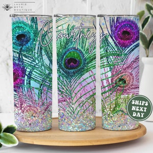 May include: Three stainless steel tumblers with a peacock feather design. The tumblers have a glittery finish and are decorated with iridescent colors. The tumblers are arranged in a row on a wooden surface.
