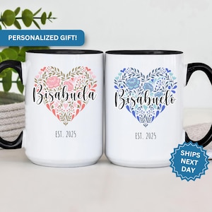 May include: Two white ceramic mugs with black handles. The mugs have a floral heart design with the words "Bisabuela Est. 2025" and "Bisabuelo Est. 2025" on them.