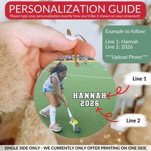May include: A round, flat, custom photo ornament featuring a field hockey player on a green field. The text "HANNAH" and "2026" is printed in white with a black outline. A red arrow points to the name and another to the year for personalization.
