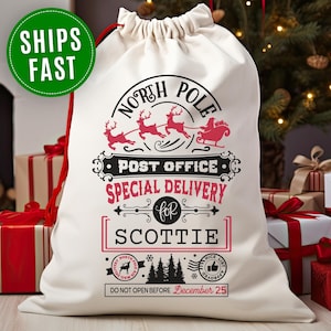 May include: A white drawstring gift bag with a red drawstring cord. The bag features a festive design with the words "North Pole Post Office Special Delivery" and the name "SCOTTIE". A green circle says "SHIPS FAST".