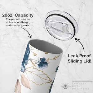 May include: A 20 ounce stainless steel tumbler with a white background and a floral design in shades of blue and gold. The tumbler has a clear, leak-proof sliding lid. The text "20oz. Capacity" and "Leak Proof Sliding Lid!" are visible in the image.