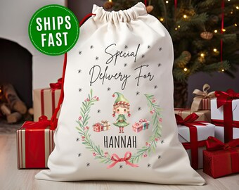 May include: A cream-colored Christmas gift bag with a red drawstring. The bag features the text "Special Delivery For HANNAH" with an illustration of an elf and gifts. A green circle with "SHIPS FAST" is in the upper left.