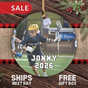 May include: A round ceramic ornament featuring a photograph of a lacrosse player in a navy blue uniform with a yellow helmet, holding a lacrosse stick. The text JONNY and 2026 is printed in white with a black outline across the center of the image.