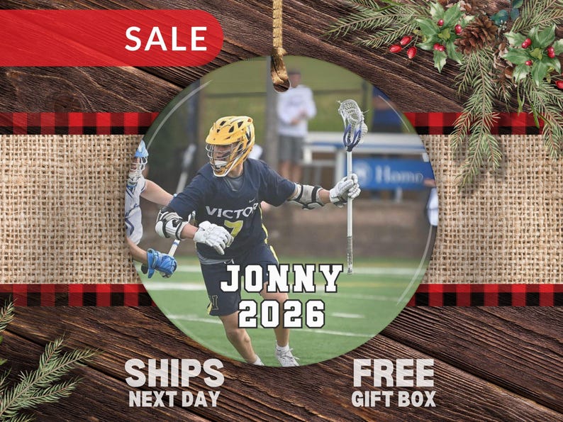 May include: A round ceramic ornament featuring a photograph of a lacrosse player in a navy blue uniform with a yellow helmet, holding a lacrosse stick. The text JONNY and 2026 is printed in white with a black outline across the center of the image.