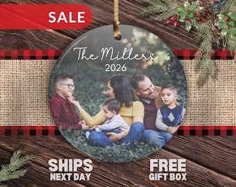 Op de afbeelding: Een ronde keramische hanger met een afgedrukte foto van een gezin van vier dat buiten zit. De tekst "The Millers" en "2026" is in witte letters bovenaan gedrukt. Een gouden lint is bevestigd aan het gat aan de bovenkant.
