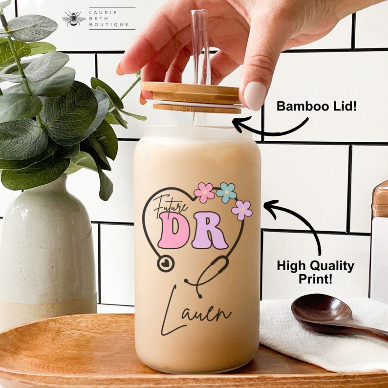 May include: A clear glass water bottle with a bamboo lid and a straw. The bottle has a design of a heart with the words "Future DR" inside and a stethoscope. The name "Lauren" is written below the heart. The bottle is sitting on a wooden surface.