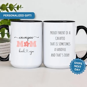 May include: White ceramic coffee mug with black rim and handle. The mug has a pink flower and the text "cavapoo MOM heidi & ajee". The other mug has black rim and handle and the text "Proud parent of a cavapoo that is sometimes a handful and that's okay".