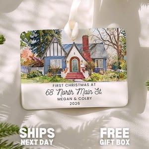 May include: A rectangular white ornament featuring a watercolor illustration of a blue house with a red chimney and steps. The text reads: FIRST CHRISTMAS AT 68 North Main St. MEGAN & COLBY 2026. It hangs from a white ribbon.