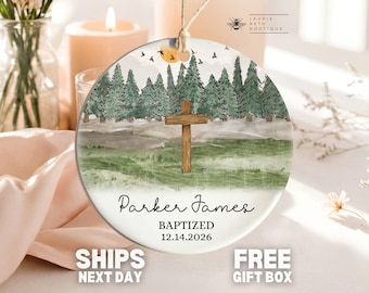 May include: A round ceramic ornament featuring a watercolor illustration of a wooden cross set against a forest of green pine trees and a sunset sky. The text reads Parker James, BAPTIZED, and 12.14.2026. It hangs from a thin twine string.