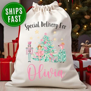 May include: Cream-colored drawstring gift bag with a festive design. The bag features a Christmas tree, a nutcracker, a ballerina, and a fox. Text on the bag reads "Special Delivery For Olivia" and "SHIPS FAST". Red drawstring closure.