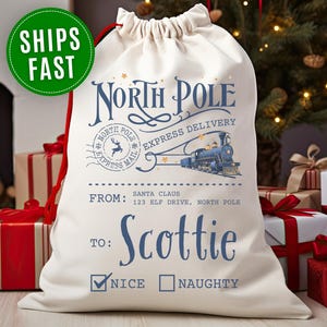 May include: Cream-colored Christmas gift bag with a drawstring closure. The bag features a blue design with the words "North Pole Express Delivery" and a train illustration. The name "Scottie" is printed below the text, with check boxes for "Nice" and "Naughty".