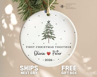 May include: A round white ceramic ornament featuring a green pine tree illustration, small black stars, and the text "FIRST CHRISTMAS TOGETHER" with the names "Olivia" and "Peter" separated by a red heart, plus the year "2026".