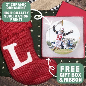 May include: A 3-inch round ceramic ornament featuring a sublimation print of a lacrosse player in a white uniform with the text MICHAEL 2026. The ornament is presented in a gift box alongside a red knitted Christmas stocking with a white letter L.