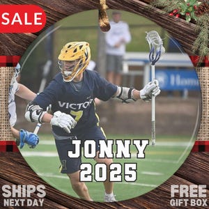 May include: Round ornament featuring a lacrosse player in a navy blue uniform with the word "VICTOR" and the number 7. The ornament has the name "JONNY 2025" in large white letters. The background shows a green field.
