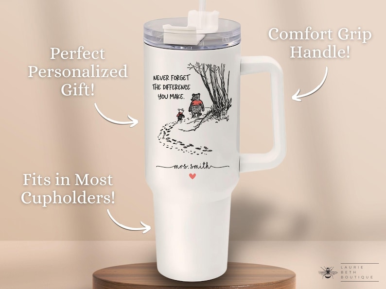 May include: White travel mug with a black and white illustration of Winnie the Pooh and Piglet walking in the woods. The mug has a black handle and a black lid with a straw. The text "Never forget the difference you make." is printed on the mug. The mug is personalized with the name "mrs. smith".