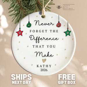 May include: A round white ceramic ornament featuring the black text Never FORGET THE Difference THAT YOU Make, a gold heart, and the name KATHY with the year 2026. The design includes small red and green star and ball ornaments and gold sparkles.