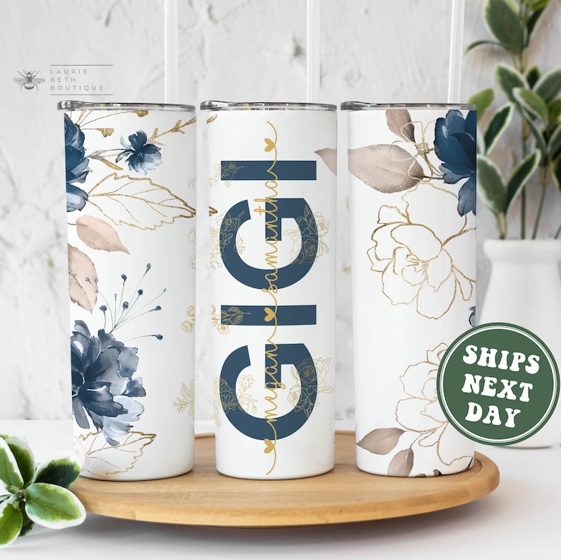 May include: Three white stainless steel tumblers with a floral design. The tumblers have a blue and gold floral pattern. The middle tumbler has the word "Gigi" written in gold letters with the text "from your favorite granddaughter" written in smaller gold letters below.