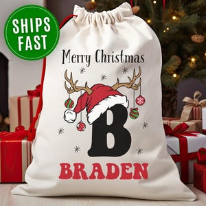 May include: A cream-colored Christmas gift bag with a red drawstring. The bag features a festive design with a Santa hat, antlers, ornaments, snowflakes, the letter "B", and the name "Braden" in red.