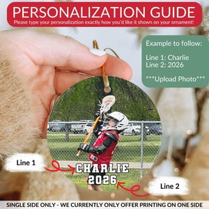 May include: A round, personalized ornament featuring a photograph of a lacrosse player in a red jersey and white helmet holding a lacrosse stick. The text 'CHARLIE' appears on line one and '2026' on line two in white block letters with black outlines.
