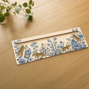 May include: A rectangular, cream-colored flat object featuring a horizontal slot at the top. The surface is decorated with a repeating pattern of orange and black tigers, blue floral blossoms, and green bamboo stalks in a detailed illustration.