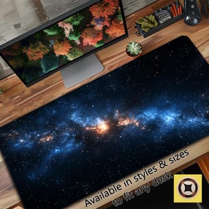 Puede incluir: Una alfombrilla de escritorio azul y negra con temática de galaxia con un diseño de nebulosa y estrellas blancas. El texto "Disponible en estilos y tamaños para adaptarse a cualquier escritorio" está impreso en la alfombrilla.