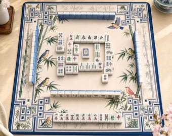 Puede incluir: Una alfombrilla de mahjong cuadrada con una superficie de color crema que presenta ilustraciones de tallos de bambú y pájaros coloridos. La alfombrilla tiene un borde azul y está dispuesta con fichas de mahjong blancas con caracteres chinos.