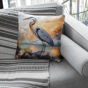 May include: A throw pillow with a colorful image of a great blue heron standing on a rock in a lake. The heron is blue-grey with a long neck and long legs. The background is a sunset sky with clouds.