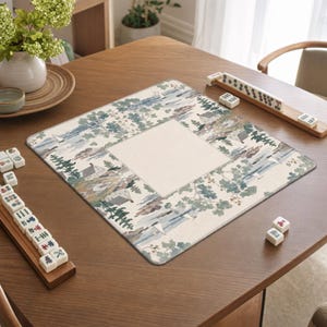 Könnte beinhalten: Eine quadratische Mahjong-Matte mit einer aufgedruckten Landschaftsszene aus Bäumen, Häusern und Segelbooten in Grün-, Blau- und Brauntönen, die ein cremefarbenes Quadrat in der Mitte umgibt, ergänzt durch hölzerne Spielsteinhalter und weiße Spielsteine.