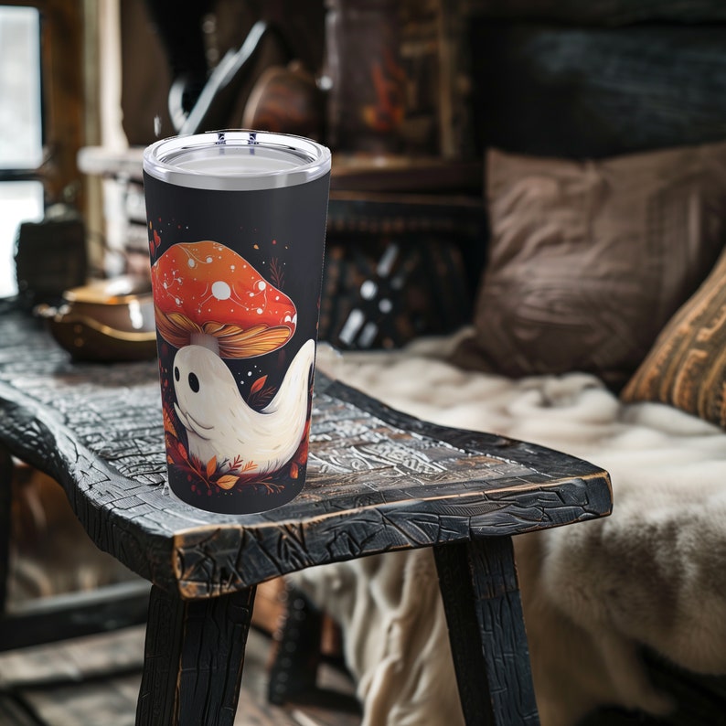 May include: A black stainless steel tumbler with a white ghost and a red and orange mushroom design. The tumbler is sitting on a wooden table.