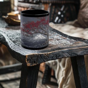 May include: Black coffee mug featuring a detailed illustration of Mount Fuji with a cherry blossom tree. The design includes shades of gray, red, and white. The mug has a black rim and a cylindrical shape.