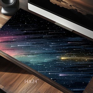 Op de afbeelding: Een rechthoekige bureaumat met een kosmisch ontwerp. De mat heeft een donkerblauwe achtergrond met strepen van wit en geel licht, en een verloop van roze en blauw. De mat meet 35,5 x 61 cm.